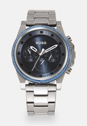 Reloj cronógrafo metálico con bisel texturizado en denim azul, esfera oscura, acentos plateados y pulsera de eslabones. Nombre de la marca "HUGO" mostrado.