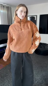 Pulôver de lã cor ferrugem com gola alta, meio fecho de correr e detalhes florais brancos nas mangas. Combinado com calças pretas de perna larga.