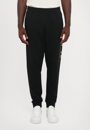 Black sweatpants with a relaxed fit, featuring an elastic waistband and cuffs, and a gold logo detail on the left side. Paired with white sneakers.