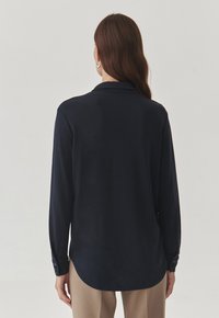 Navy blauw langemouw shirt met een kraag, met een gladde textuur en knoopmanchetten, en loopt naar een afgeronde zoom aan de achterkant.