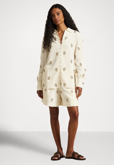 Svetlo béžový dvojdielny outfit s košeľou s golierom a zodpovedajúcimi šortkami, ozdobený výšivkou a detailmi na gombíky; voľný strih a ľanový materiál.