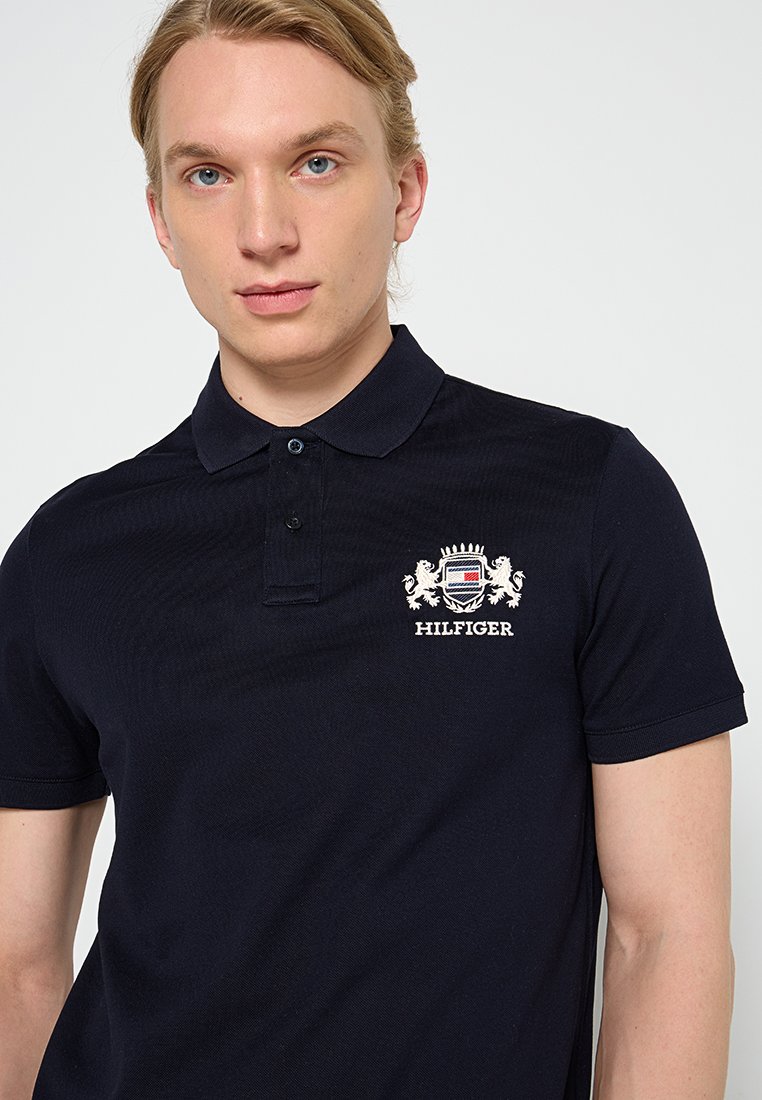 Jeune homme aux cheveux blonds portant un polo bleu marine avec un blason brodé et le texte "HILFIGER" sur la poitrine gauche.
