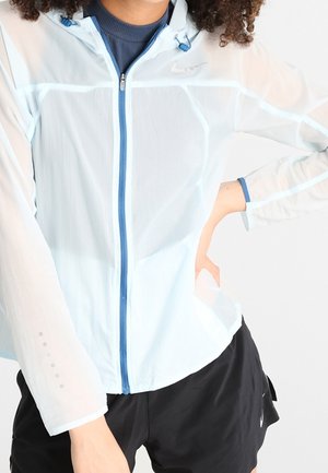 Personne portant une veste zippée bleu clair avec capuche et un short de sport noir, se tenant avec une main sur la hanche.