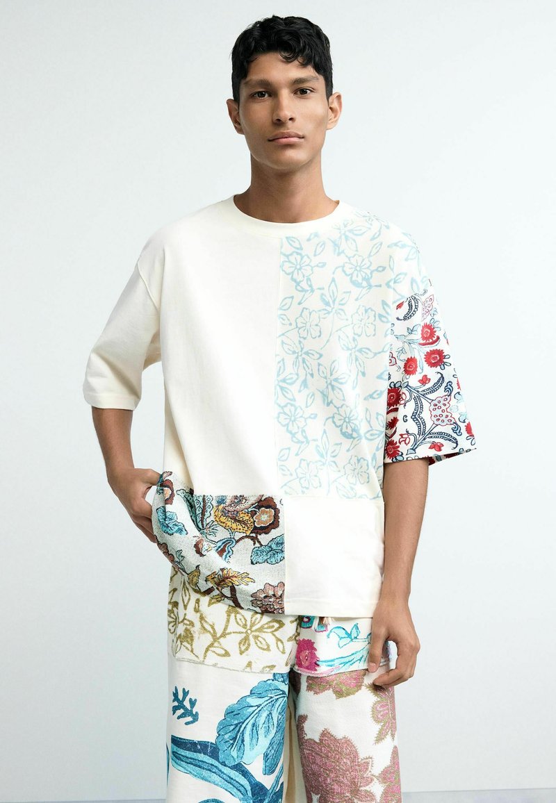 Young man wearing oversized patchwork outfit with mixed floral patterns in blue, red, green, and beige tones, standing against plain white background.