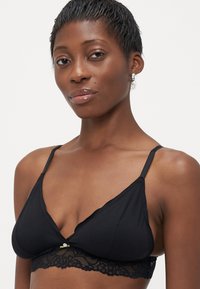 Vrouw met kort haar, gekleed in een zwarte bralette met kant en kleine zilveren oorbellen, tegen een effen witte achtergrond.