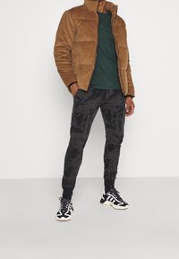 Bruine corduroy jas met hoge kraag, groene shirt en zwarte, geblokte joggers. G gedragen met zwarte sneakers met witte accenten.