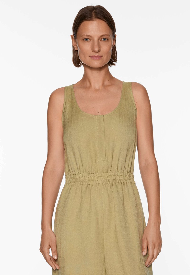 Frau mit kurzen braunen Haaren, die einen ärmellosen, hellolivenfarbenen Jumpsuit mit elastischem Bund trägt, steht vor einem weißen Hintergrund.