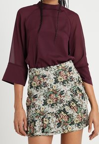 Bordeaux lange mouwen blouse met hoge hals, gecombineerd met een meerkleurige bloemenrok met ruffles in een groen, blauw en beige patroon.