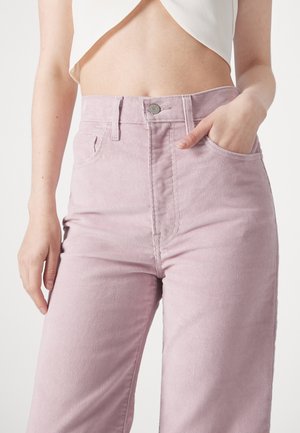 Femme portant un pantalon en velours côtelé violet clair taille haute avec un crop top blanc, une main dans la poche, sur un fond uni.