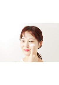 Feuchtigkeitsspendende Creme, die mit einem Finger auf einer Wange aufgetragen wird. Weiße cremige Textur auf makelloser Haut, hellem Hintergrund und natürlichem Make-up.