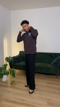 Brun hættetrøje med lynlås og hvidt logo, sorte bukser og brune sko. Værelset har grøn sofa og plante, med trægulv og lys.