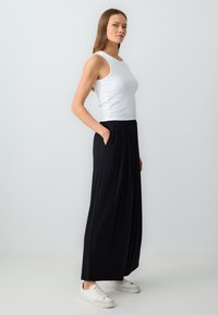Débardeur sans manches blanc associé à une jupe maxi noire fluide à taille haute avec poches. Le mannequin porte des baskets blanches.
