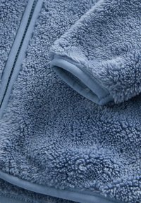 Fuzzy blauwe fleece stof met een gestructureerd oppervlak, voorzien van een zachte zoom en ritsdetail, wat zorgt voor een zachte en gezellige uitstraling.