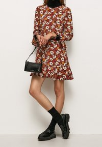 Kvinna klädd i en röd blommig klänning över en svart turtleneck, svarta strumpor, kraftiga svarta loafers och håller en liten svart handväska.