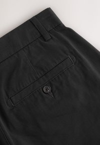 Primer plano del bolsillo trasero de pantalones negros con cierre de botón y presilla para cinturón en tela lisa.