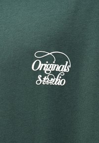 Σκούρο πράσινο βαμβακερό ύφασμα με λευκό κεντητό λογότυπο που γράφει "Originals Studio", με καλλιγραφικό σχέδιο και διακοσμητικές λεπτομέρειες.