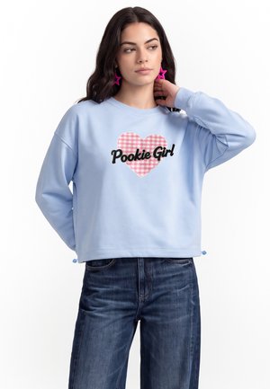 Donna con capelli scuri indossa una felpa blu con un cuore a quadretti rosa e il testo "Pookie Girl", abbinata a jeans blu scuro e orecchini a forma di stella rosa.