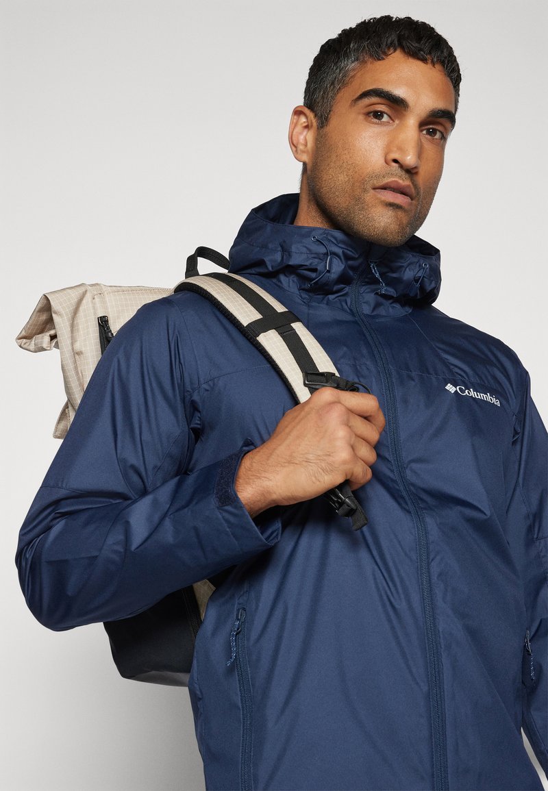 Marineblaue wasserdichte Jacke mit Reißverschluss-Taschen und hohem Kragen; getragen mit einem beigen und schwarzen Rucksack mit gepolsterten Trägern.