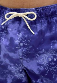 Lila batik mintás fürdőnadrág fehér nap grafikákkal. Rugalmas derékpánttal és műanyag végekkel ellátott húzózsinórral rendelkezik.