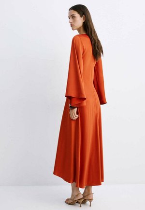 Vrouw met lang bruin haar, zijwaarts kijkend, draagt een lange oranje jurk met wijde mouwen en beige sandalen met hakken.