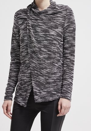 Žena v černobílém pleteném svetru s volným límcem a černých přiléhavých kalhotách stojí na jednobarevném pozadí.