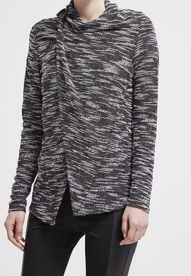 Femme portant un pull en maille chiné noir et blanc avec un col bénitier drapé et un pantalon noir ajusté, debout devant un fond uni.