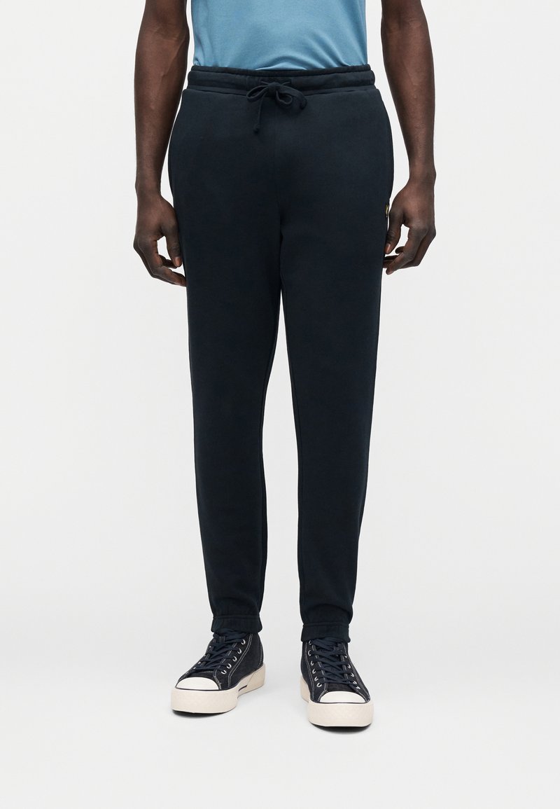 Muž nosiaci čierne teplákové nohavice s elastickými lemami, modré tričko zastrčené dovnútra a čierne vysoké tenisky s bielymi podrážkami.