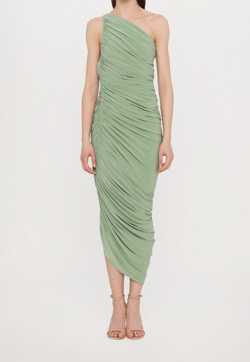 Femme portant une robe midi vert clair asymétrique à une épaule avec fronces et des talons nus à brides aux chevilles, debout devant un fond uni.