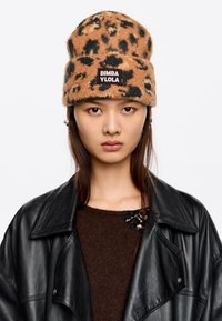 Gorro de punto con estampado de leopardo marr�ón y negro, con un diseño de doblez y un parche de marca. El modelo lleva una chaqueta de cuero negra y un suéter marrón.