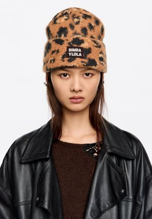 Gorro de punto con estampado de leopardo marrón y negro, con un diseño de doblez y un parche de marca. El modelo lleva una chaqueta de cuero negra y un suéter marrón.