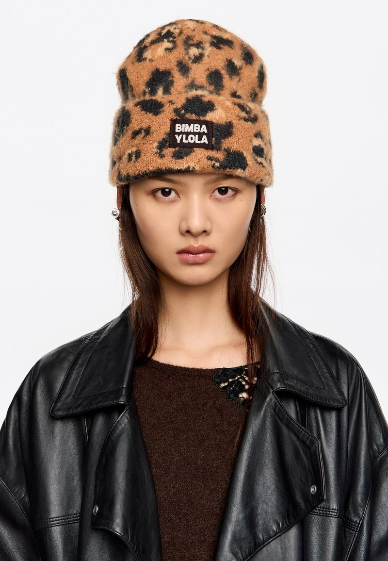 Gorro de punto con estampado de leopardo marrón y negro, con un diseño de doblez y un parche de marca. El modelo lleva una chaqueta de cuero negra y un suéter marrón.