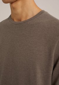 Sweatshirt texturat de culoare maro, cu guler rotund, având un model moale și ușor ridicat. Vedere detaliată a zonei pieptului și umărului.