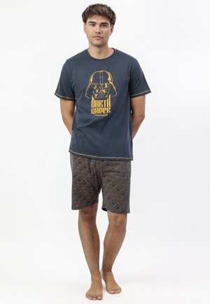 Joven de pie descalzo, vistiendo una camiseta oscura de Star Wars con Darth Vader y pantalones cortos con estampado, contra un fondo blanco.