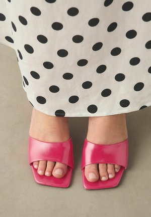 Fødder iført lyserøde sandaler med firkantet tå under en hvid nederdel med sorte polkaprikker, stående på en beige gulv.