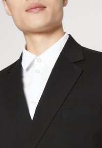 Chaqueta de traje negra con solapas tipo muesca, un bolsillo en el pecho, combinada con una camisa blanca de vestir que presenta un cuello con botones y placket.