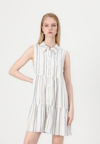 Robe sans manches à boutons en blanc avec des rayures verticales bleues et noires, dotée d'un col et d'un design en niveaux. Tissu léger avec une texture lisse.