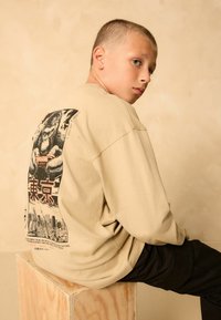 Beige méretű pulóver, hátulján egy grafikával, amelyen egy gorilla játékkonzolt tart, sötét nadrággal párosítva.