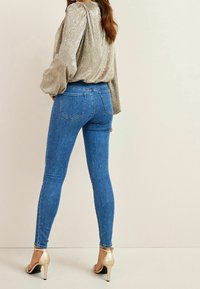 Jean skinny bleu à taille haute, orné de petites rivets décoratifs. Accompagné d'un haut long à manches en argent texturé et de talons aiguilles beiges.