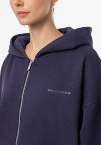 Námornícka zipsová mikina vyrobená z tkaniny s hladkou textúrou. Má veľkú kapucňu a malé, diskrétne logo "PEGADOR" v sivej farbe na hrudi.