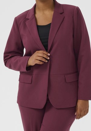 Osoba ubrana w ciemnofioletową marynarkę i spodnie, zapinająca pojedynczy guzik marynarki na czarnej bluzce.