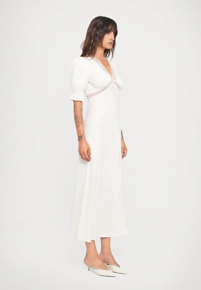 Witte lange jurk met puff mouwen en kantafwerking bij de halslijn, met een getailleerde corsage en een uitlopende rok. G gedragen met witte muiltjes.