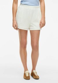 Shorts blancs texturés avec une taille élastique, offrant une coupe ample et des motifs subtils. Associés à des baskets marron et un haut bleu clair.