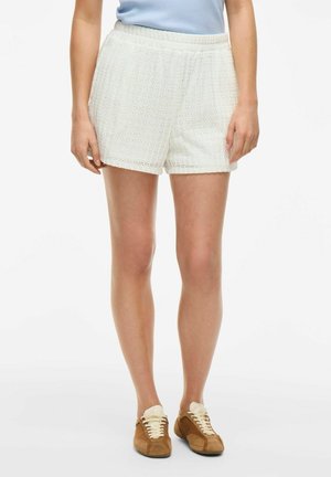 Pantaloni corti bianchi con una cintura elastica, caratterizzati da una vestibilità ampia e motivi sottili. Abbinati a sneakers marroni e a una maglietta blu chiaro.
