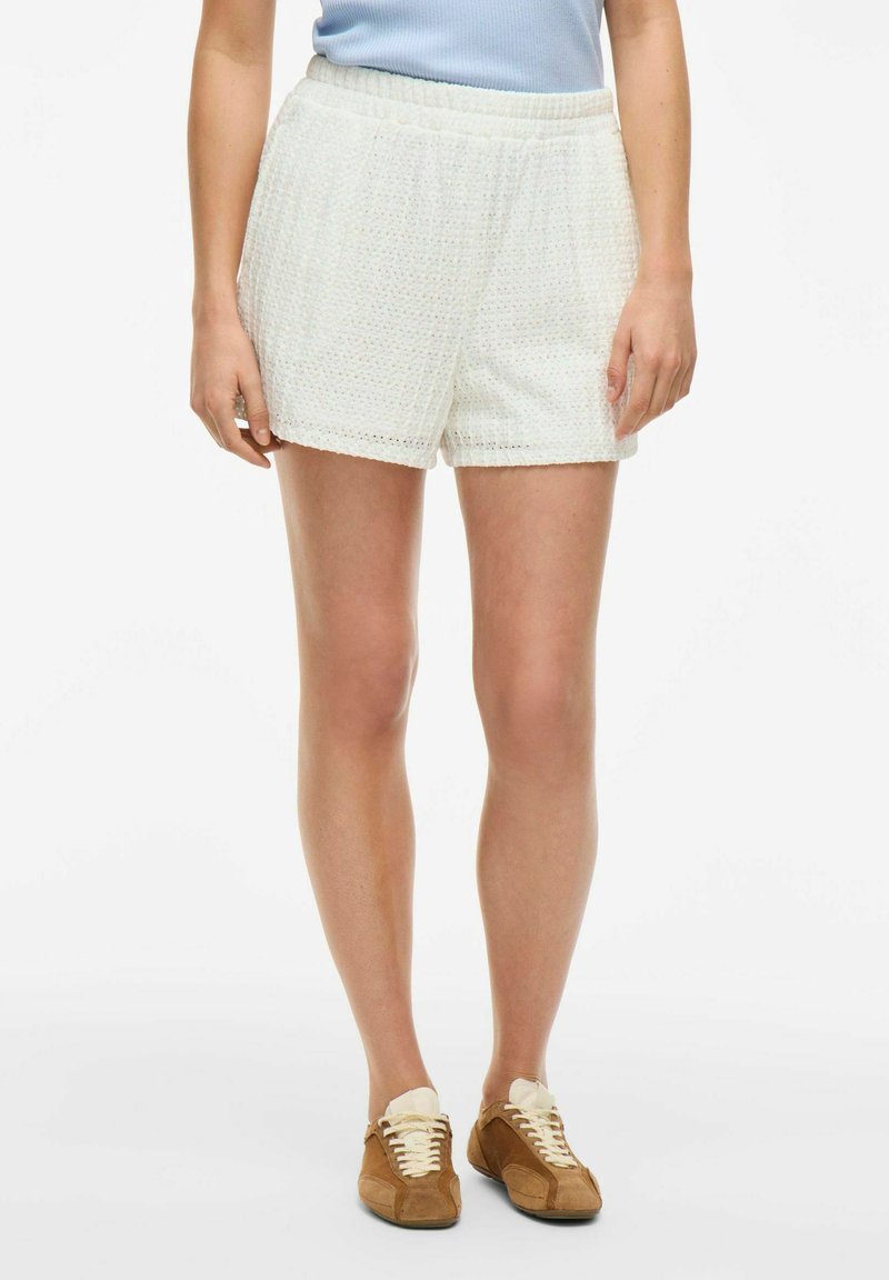 Shorts blancs texturés avec une taille élastique, offrant une coupe ample et des motifs subtils. Associés à des baskets marron et un haut bleu clair.