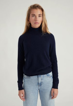 Marineblauer Rollkragenpullover aus weichem, strukturiertem Stoff; taillierte Passform mit langen Ärmeln, kombiniert mit hellblauen, hochgeschnittenen Jeans.