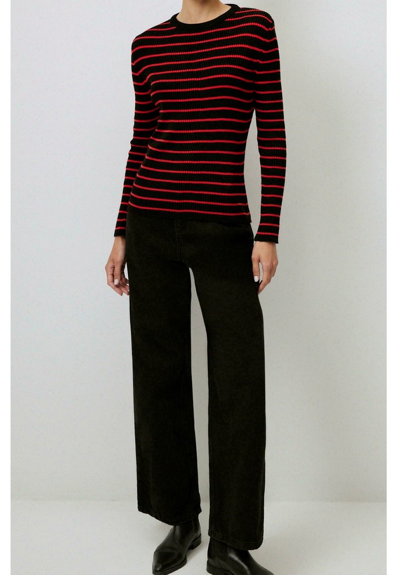 Personne portant un pull ajusté à rayures horizontales noires et rouges, avec un pantalon large noir et des bottines noires.