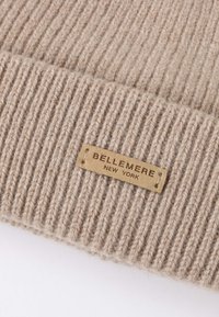 Beige ribbed knit hat featuring a rectangular leather label embossed with "BELLEMERE NEW YORK." Soft, textured material.