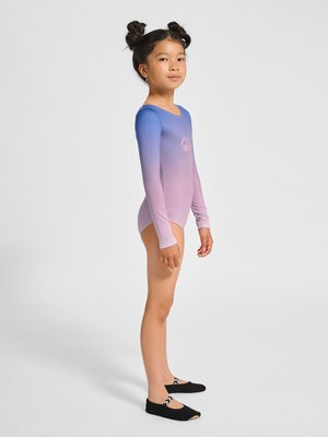 Młoda dziewczyna z kokami, ubrana w długi rękawowy kostium gimnastyczny w kolorze przejścia od niebieskiego do różowego oraz czarne baletki, stojąca bokiem na jednolitym tle.