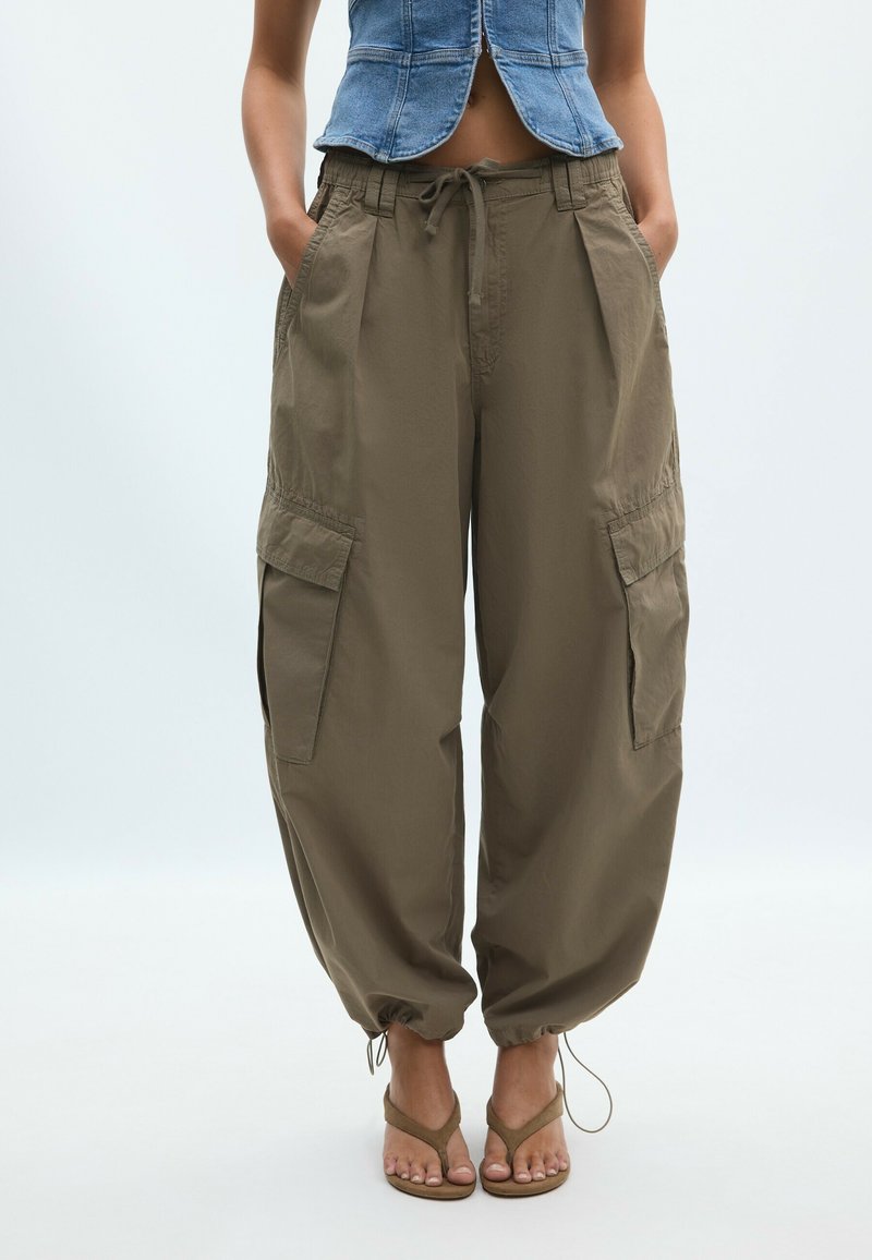 Femme portant un pantalon cargo marron large avec de grandes poches latérales et des ourlets à cordon, associé à un top en denim sans manches et des sandales.