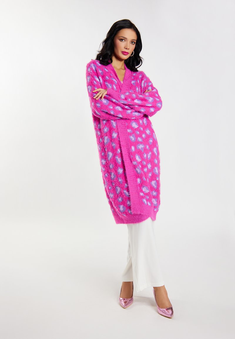 Cárdigan de felpa rosa con lunares azules, de frente abierto, mangas largas y un corte relajado, combinado con pantalones blancos de campana y tacones rosa brillantes.